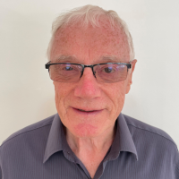 An older man with short white hair and glasses smiles slightly. He is wearing a gray collared shirt and is standing against a plain white background.