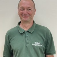 A man smiling at the camera, wearing a green polo shirt with the text "Hellingly Parish Council" embroidered on it, standing against a plain light-colored wall.