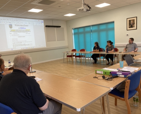 IMG_5302 A group of people sit around tables in a meeting room, looking at a document projected onto a screen. Laptops, files, and papers are on the tables. Four people sit together on one side, others are spaced around the room.