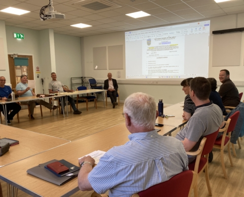 IMG_5306 A group of people sit around tables arranged in a U-shape in a meeting room, facing a large screen displaying a document. Some people are engaged in conversation; notebooks and water bottles are on the tables.