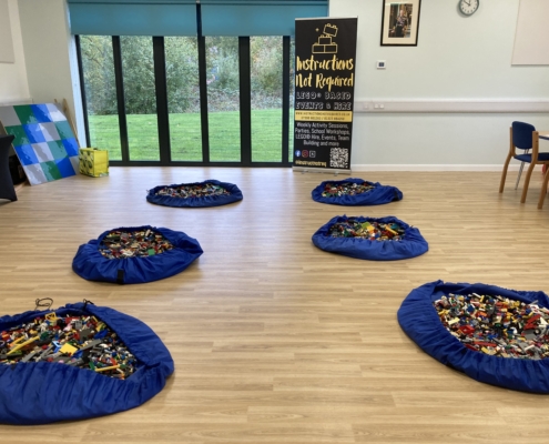 IMG_6189 A room with five blue mats filled with LEGO bricks on a wooden floor, several chairs along the wall, and a banner that says "Instructions Not Required." Large windows show greenery outside.