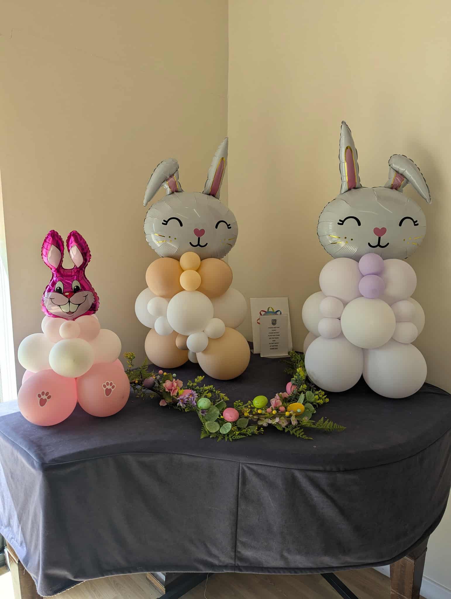 Three bunny-shaped balloon sculptures, two large and pastel-coloured, one small and pink, are displayed on a table with a spring-themed floral garland and a small sign in the background.