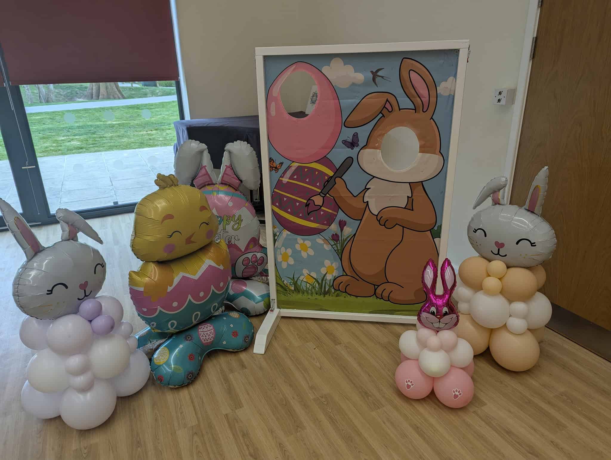A festive Easter scene with bunny and chick balloons around a standee featuring a cartoon bunny holding an egg and a balloon, with cut-outs for faces. The setup is indoors near a window and a wooden door.