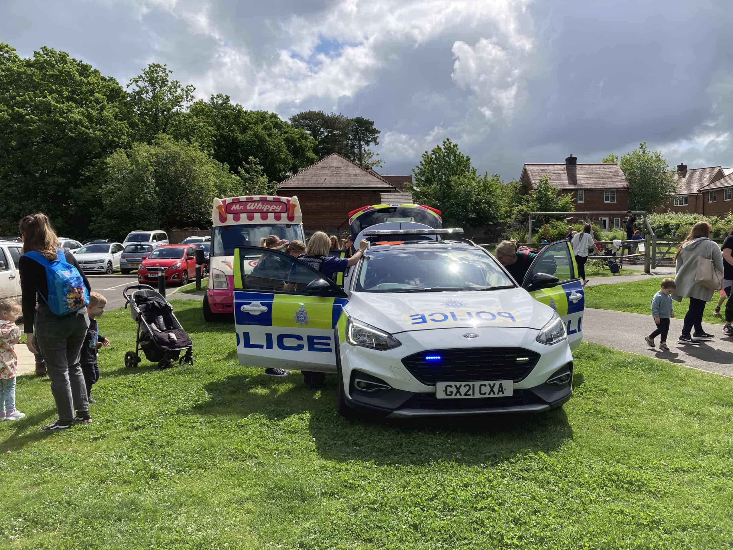 A police car with its lights on is parked on grass at a community event. People, including children, are gathered round it and an ice cream van is visible in the background, with houses and trees behind.