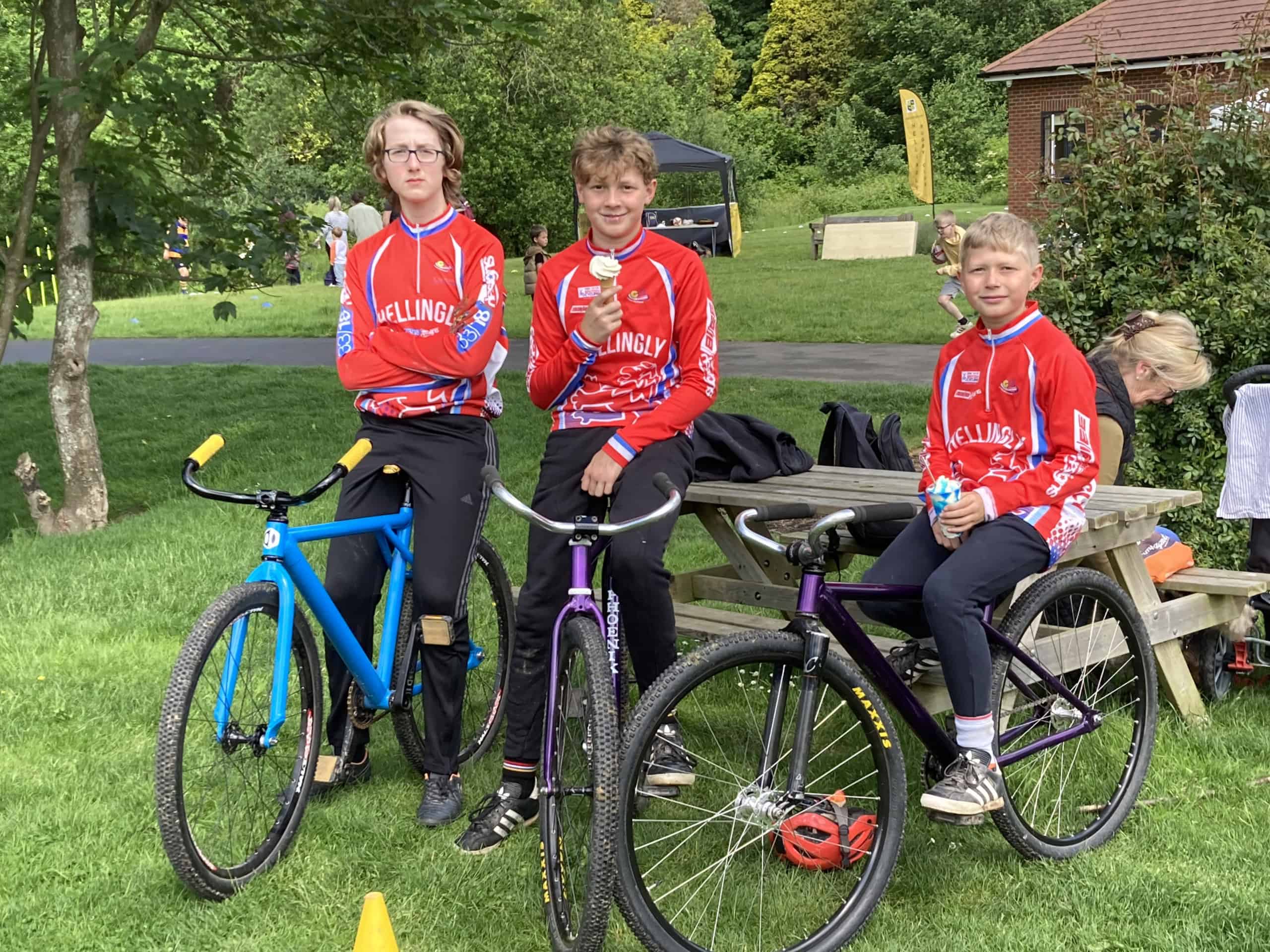 Three boys in red cycling jerseys sit and stand beside bikes on a grassy area. Two sit on a wooden picnic table, one stands holding a blue bike. Trees and a brick building are visible in the background.