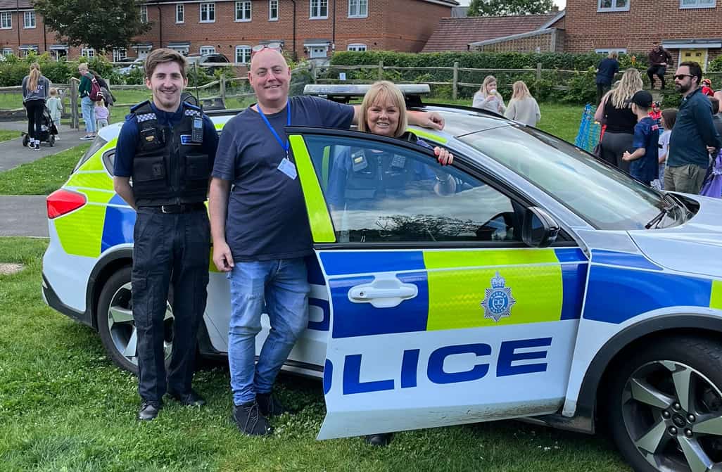 A police officer stands next to a man and a woman who are smiling by an open door of a parked police car on grass, with people and houses visible in the background.