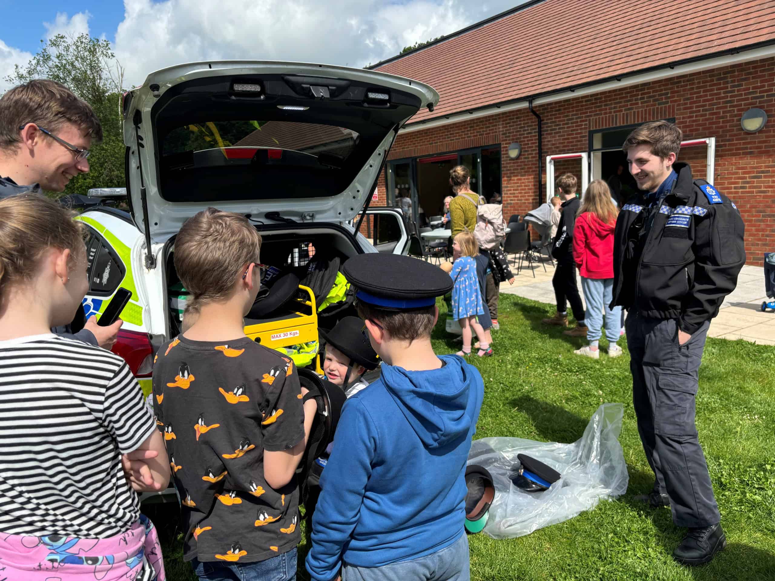 A group of children and adults gather around an open police car boot on a sunny day. A police officer stands nearby, smiling, while some kids try on police hats. The scene takes place outside a brick building.