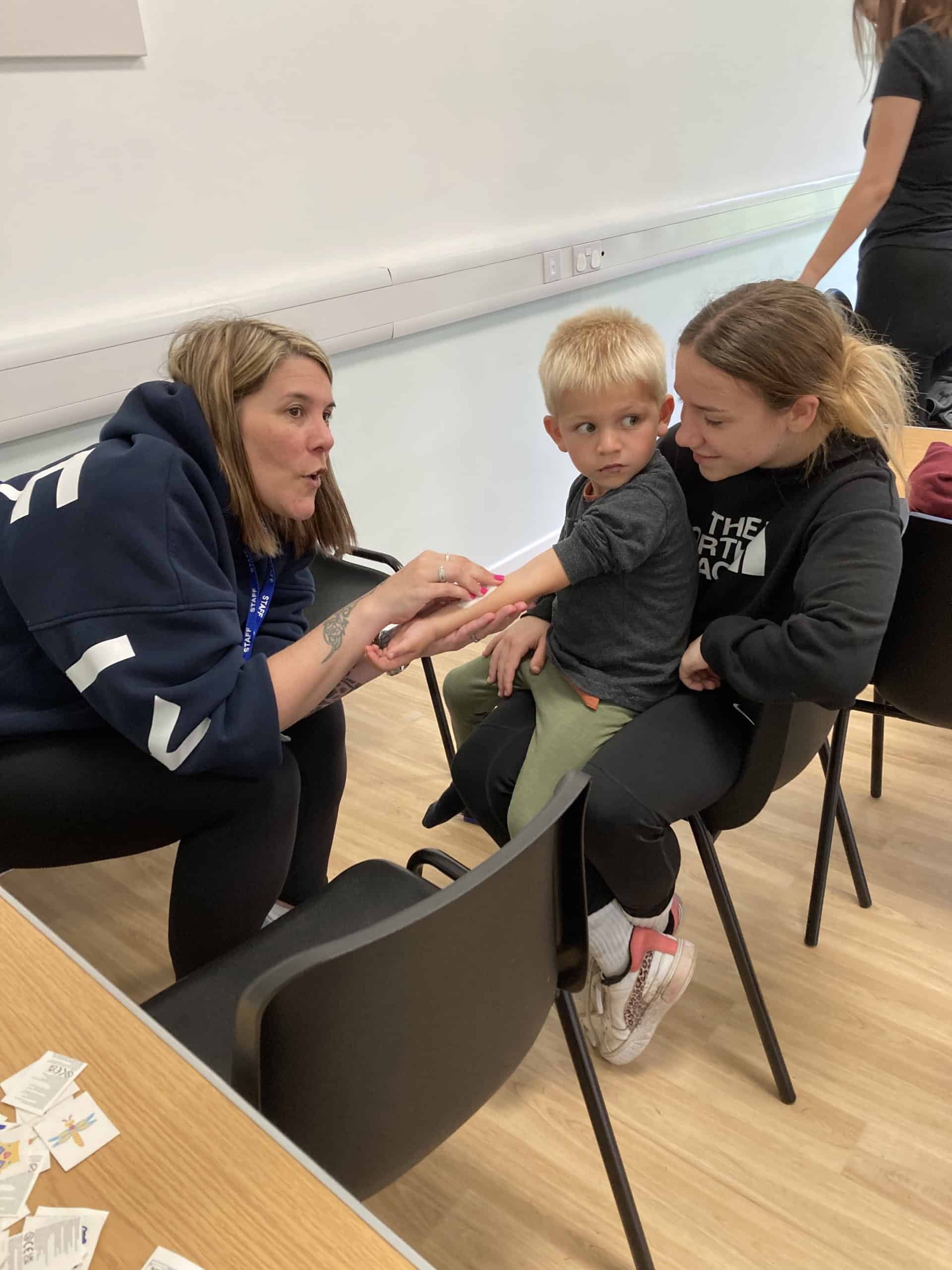 A woman applies a plaster to a young boy’s arm while he sits on a woman’s lap. They are indoors, sitting on black chairs, and the boy looks slightly unsure.