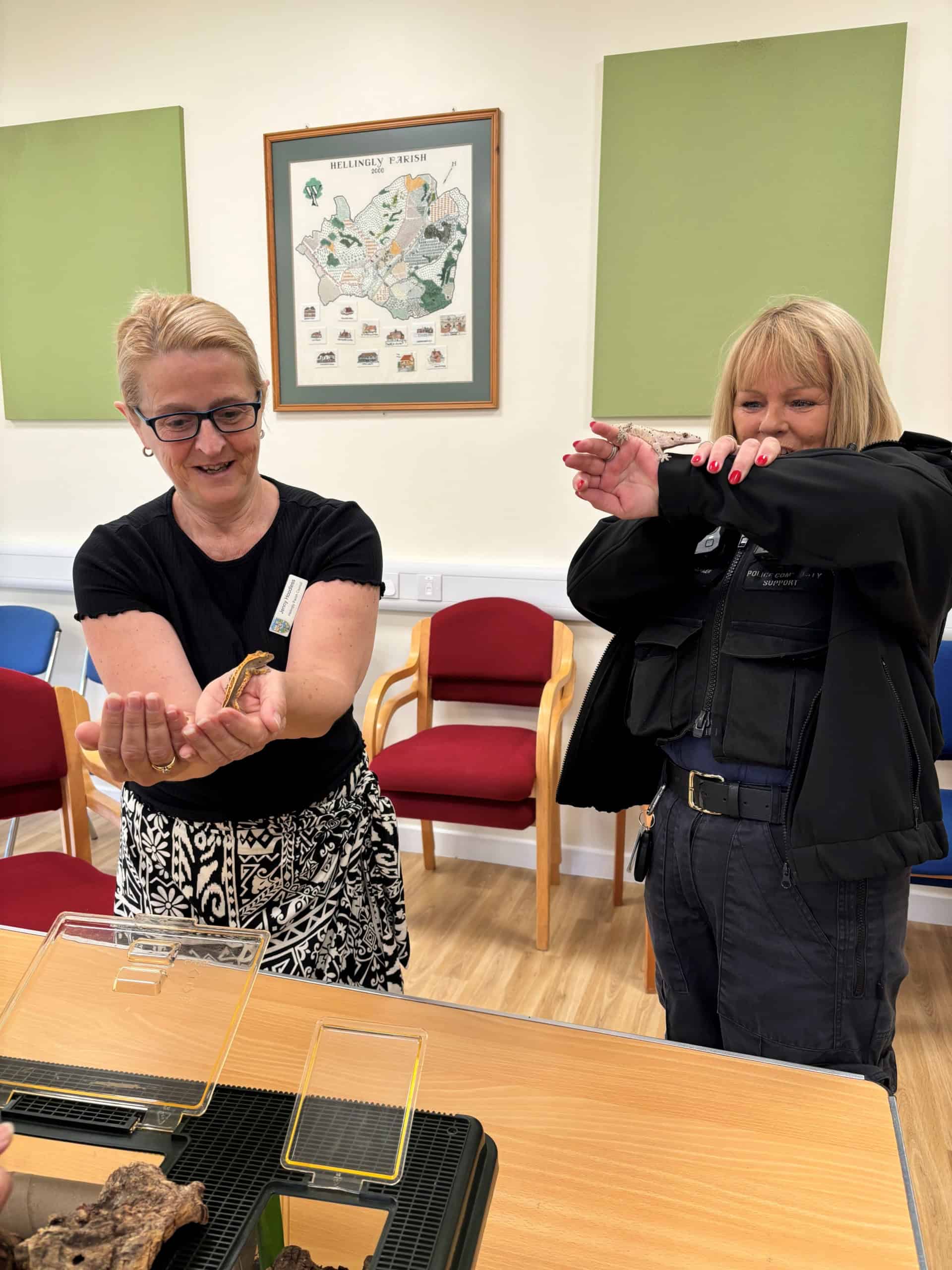 Two women stand indoors; one smiles while holding a butterfly on her hand, and the other, in a dark jacket, looks amused or hesitant, holding her arms up. Chairs and a wall map are visible in the background.