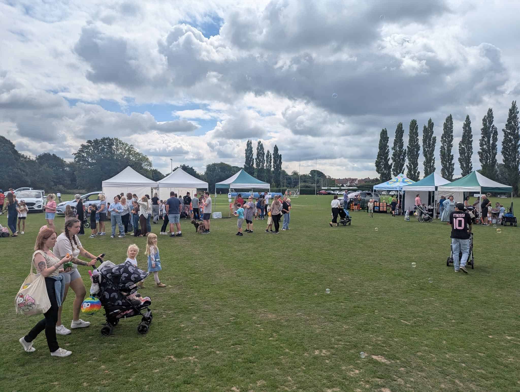 A community outdoor event with families and children walking and playing on a grassy field, several white and green marquees in the background, and a partly cloudy sky above.