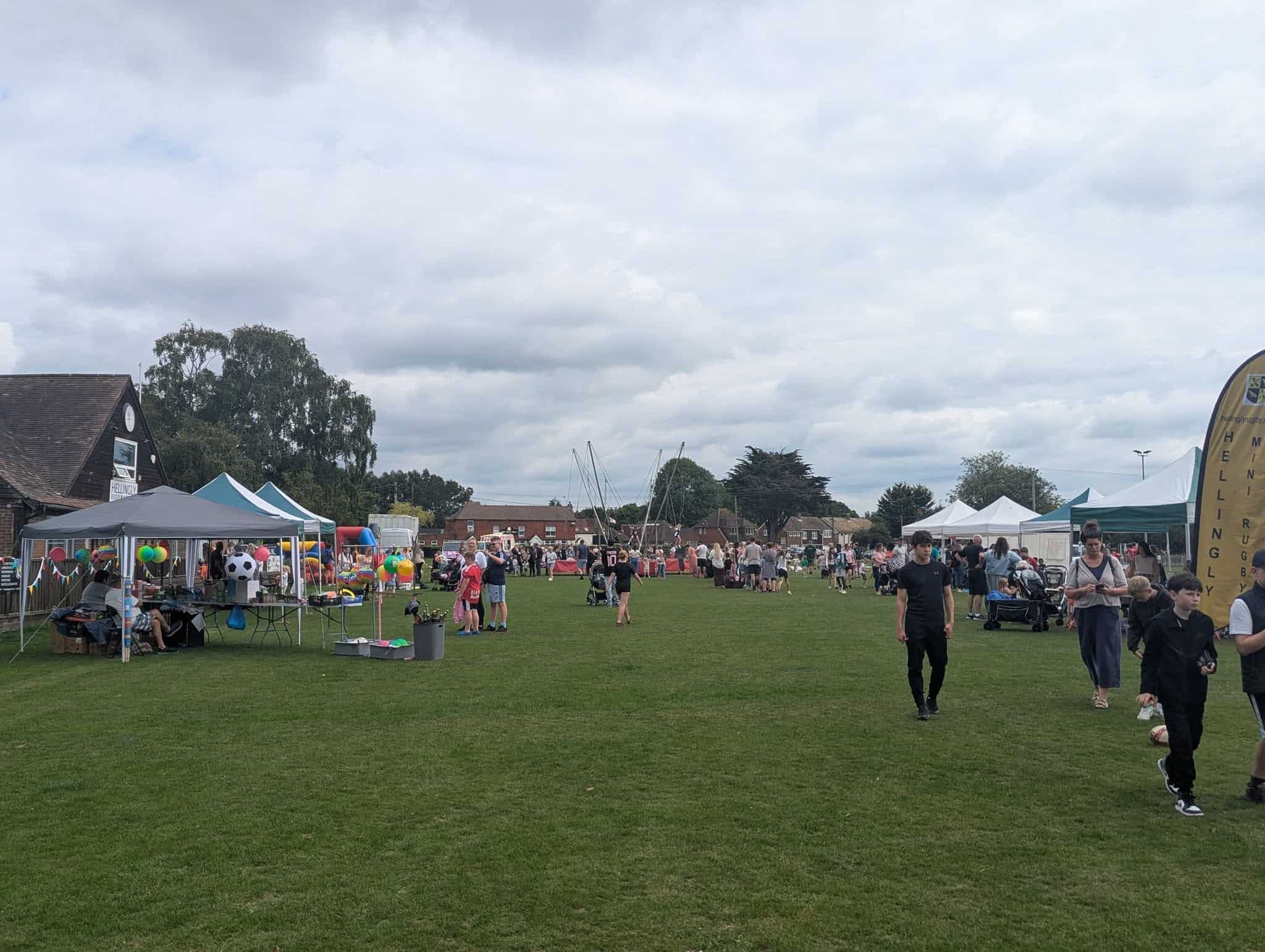 A community fete on a grassy field with people walking, marquees and stalls set up, and balloons visible. The sky is cloudy, and trees and buildings are in the background.