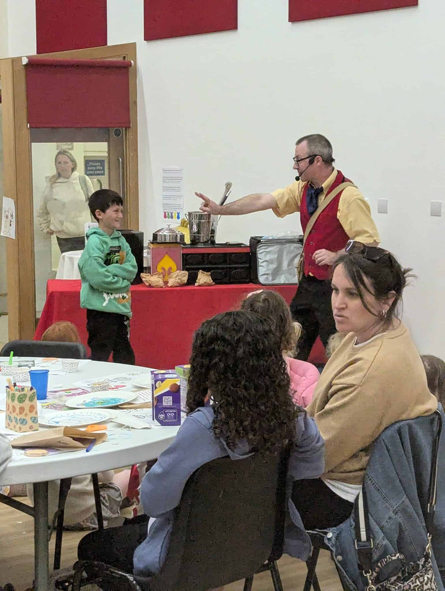A performer in a red waistcoat gestures while speaking to a boy in a green hoodie at the front of a room. Several children sit at tables watching, with arts and crafts materials on the tables. A few adults are also present.