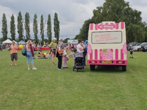 People gather around a pink and white Mr Whippy ice cream van in a park during an outdoor event, with families, children, and various activities visible in the background.