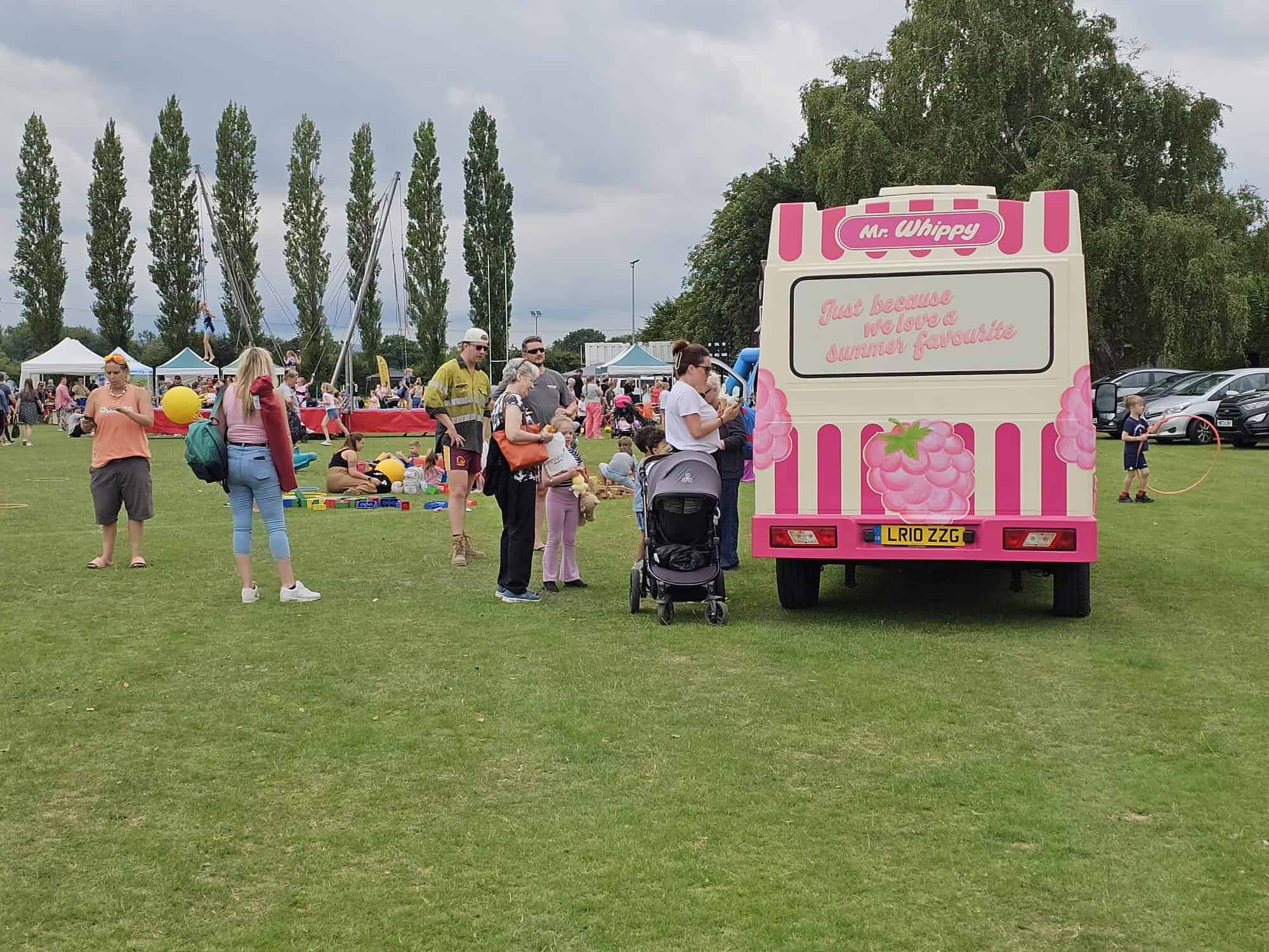 People gather around a pink and white Mr Whippy ice cream van in a park during an outdoor event, with families, children, and various activities visible in the background.