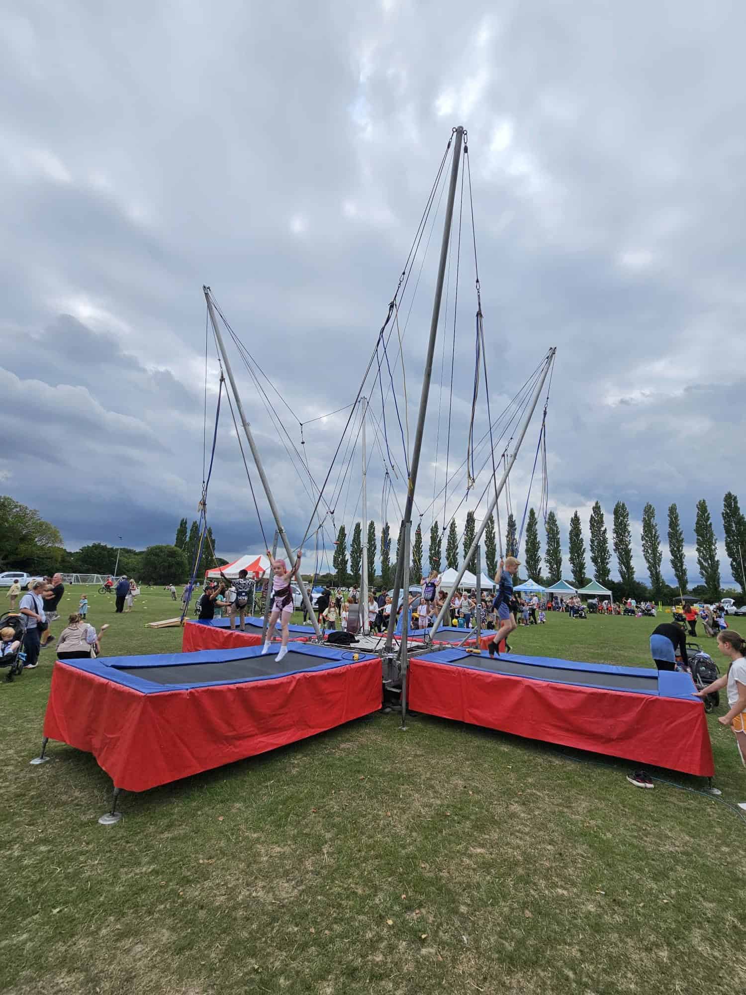 A bungee trampoline set-up at an outdoor event, with people gathered around on a grassy field. Tents and tall trees are in the background under a cloudy sky.