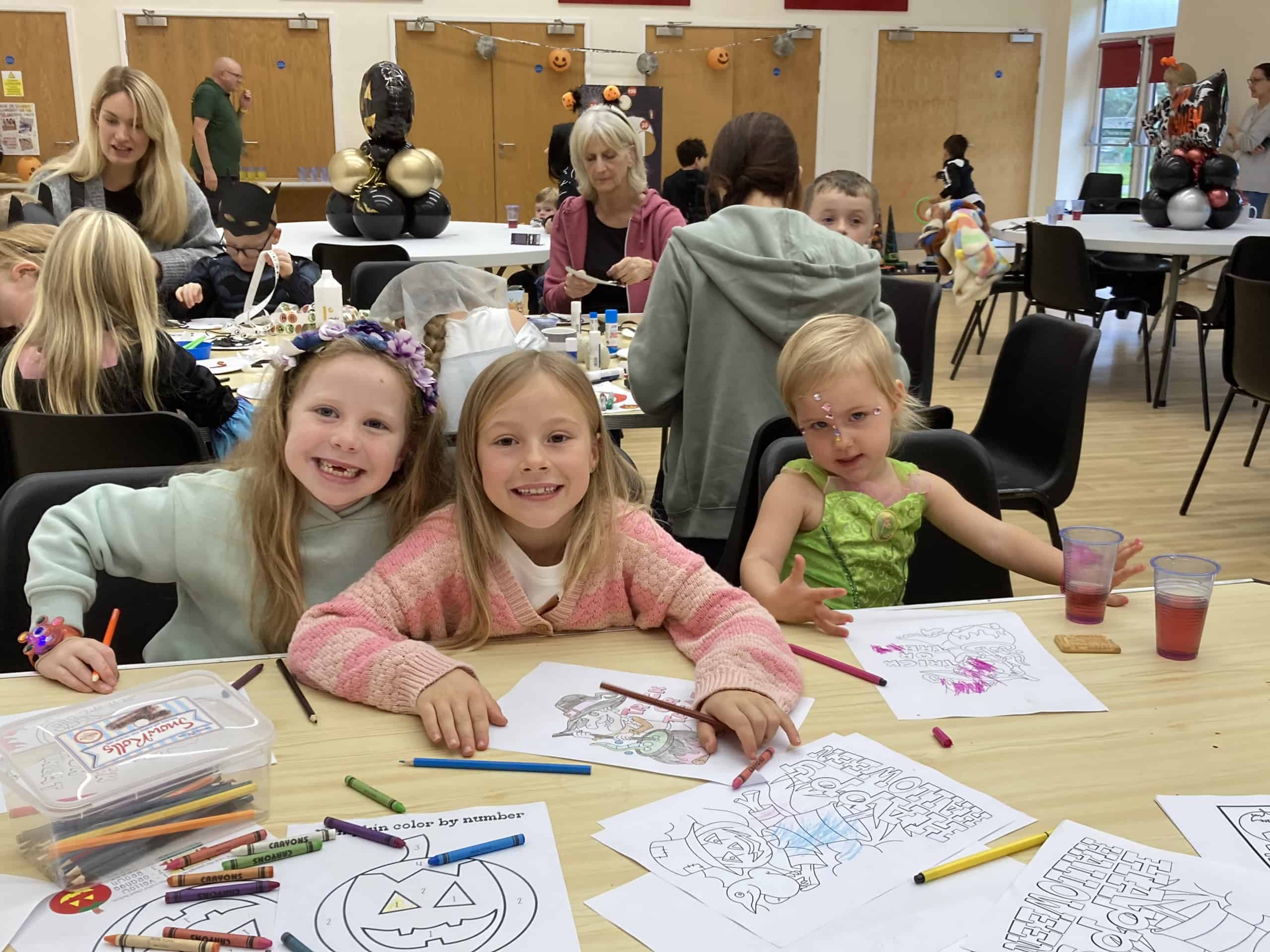 Three young girls sit at a table, smiling and colouring Hallowe’en-themed pages with coloured pencils. Other children and adults are busy with crafts in a decorated room with festive balloons and pumpkins.
