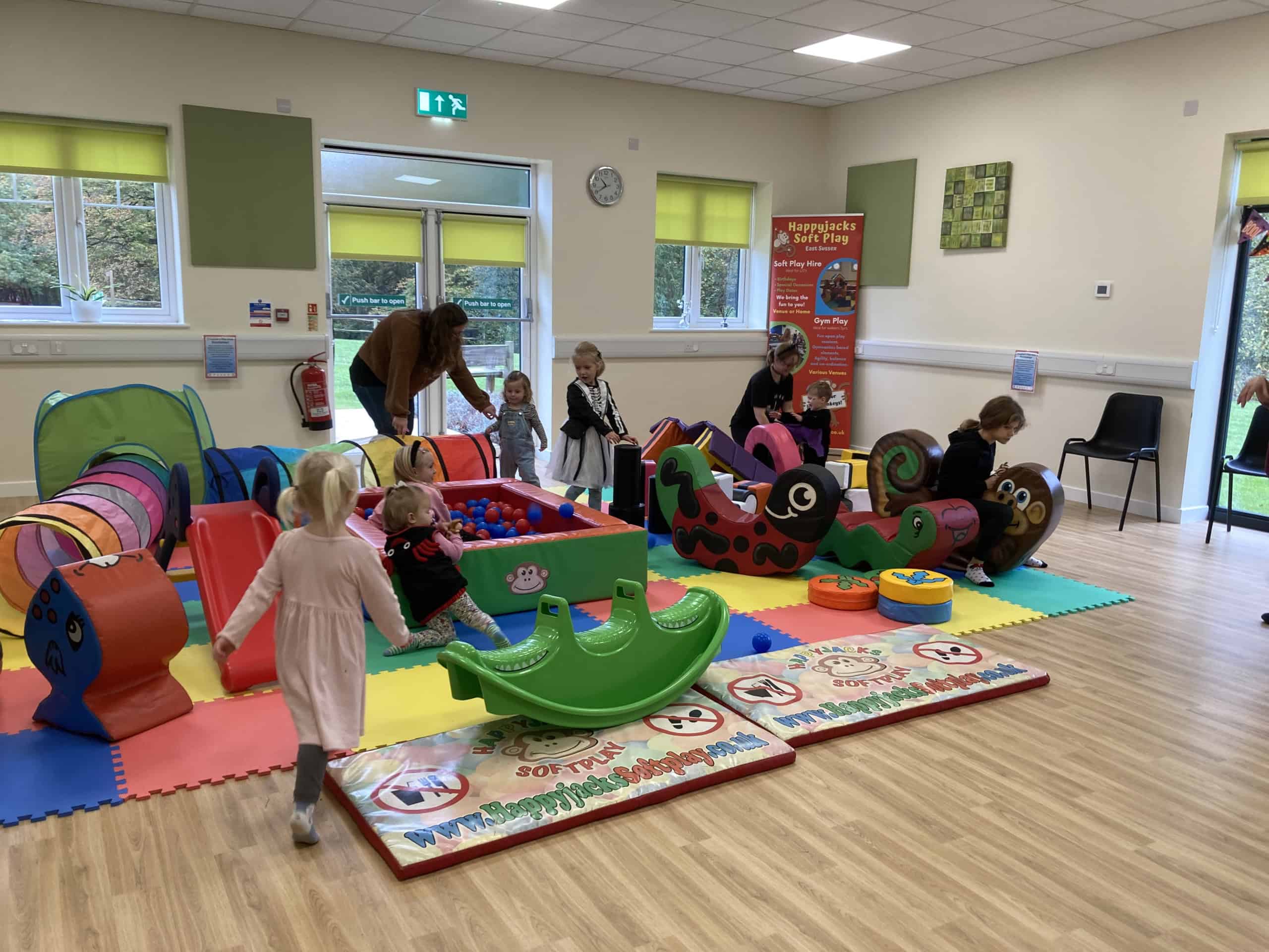Children play on colourful soft play equipment in a bright indoor playroom with foam mats, slides, and a ball pit. Adults supervise nearby. The room has large windows, light walls, and wooden floors.