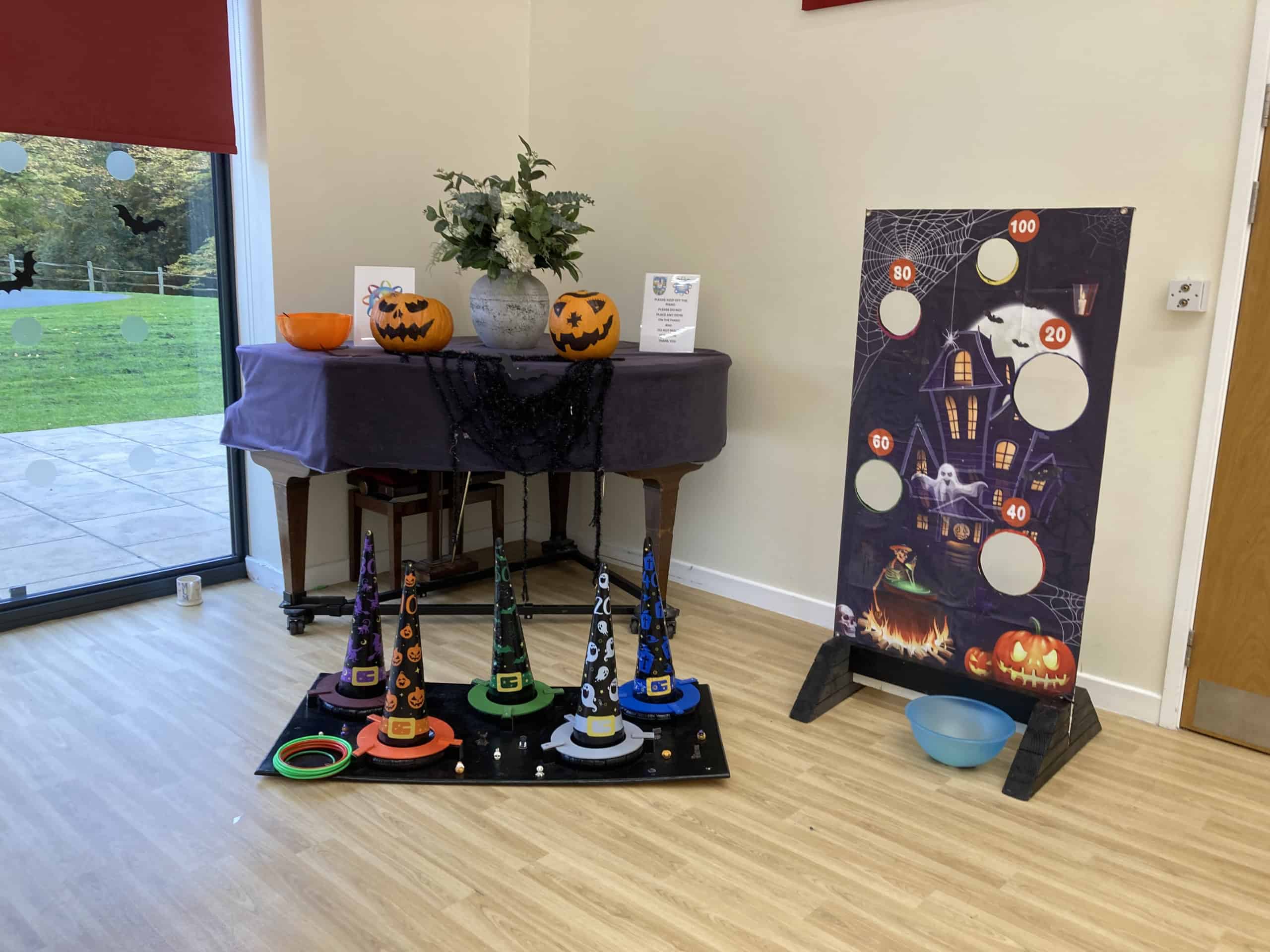 A Halloween-themed game setup indoors features pumpkin decorations, witch hats with rings for a ring toss game, and a spooky scoreboard with holes and point values. A table and a window with a garden view are in the background.