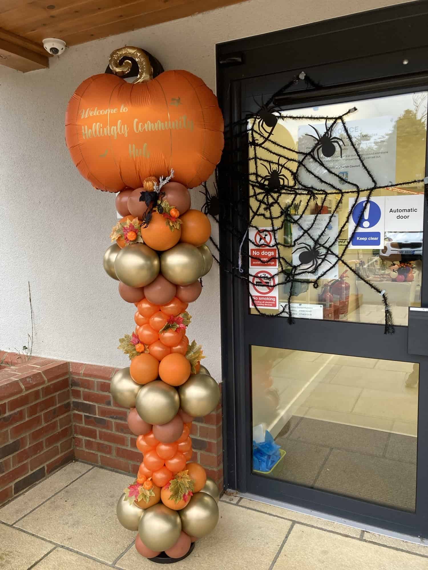 A tall balloon decoration with orange and gold balloons, autumn leaves, and a large pumpkin-shaped balloon that says “Welcome to Holliday Community Hub” stands by a door decorated with a black spider’s web.