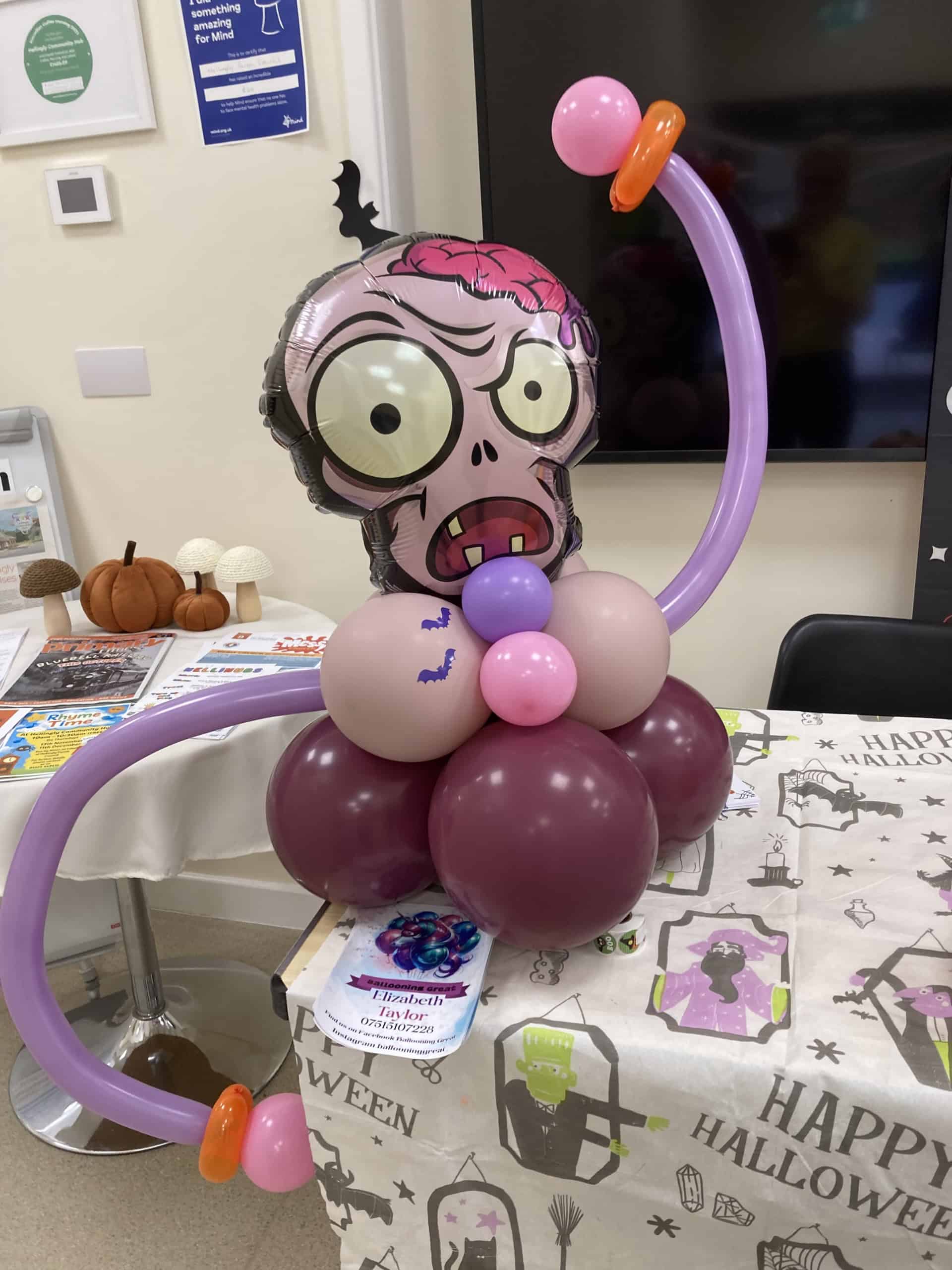 A Halloween balloon sculpture of a cartoonish zombie head with big eyes and exposed brain, surrounded by purple and pink balloons, sits on a decorated table with a "Happy Halloween" tablecloth.