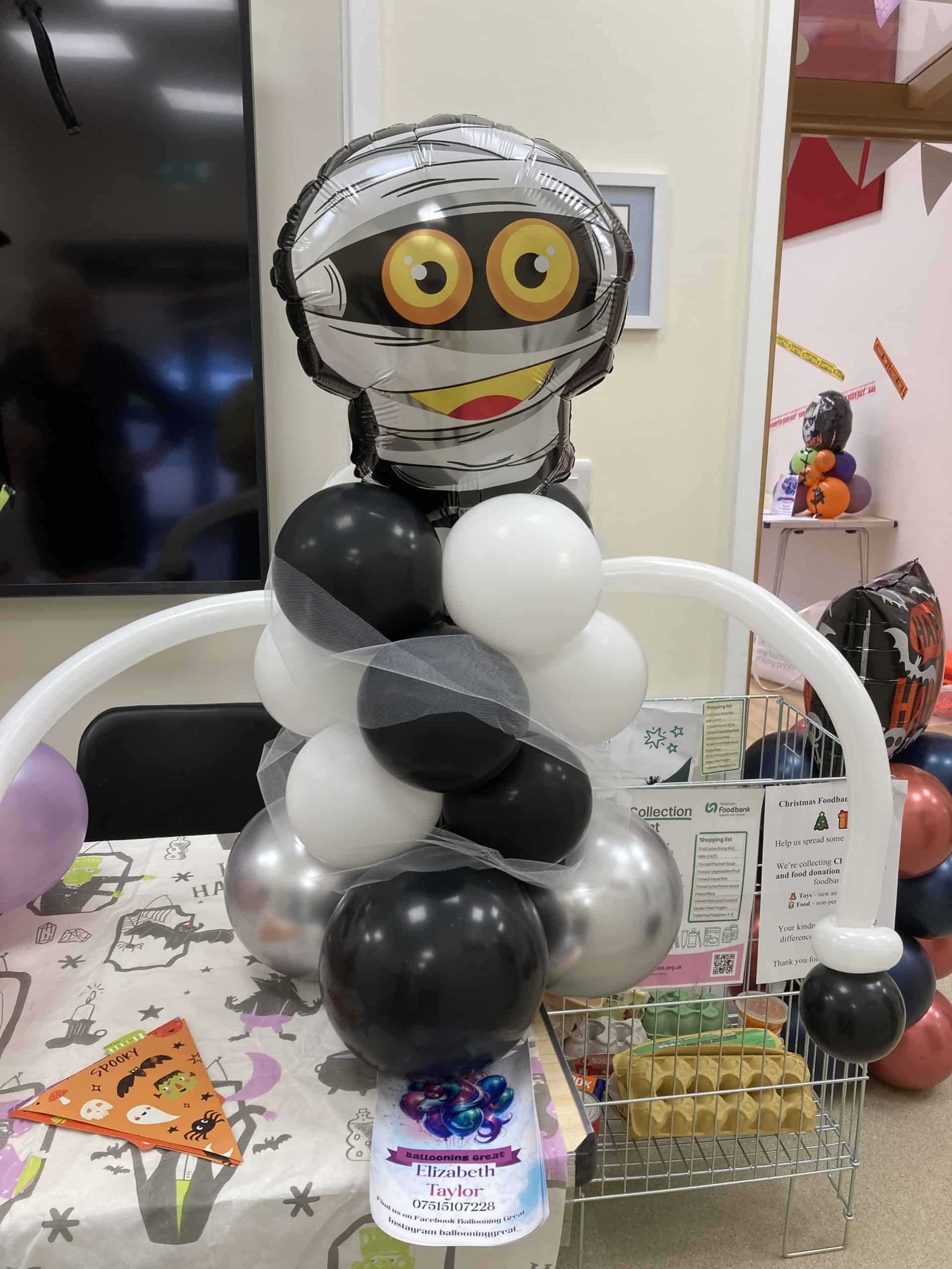 A robot figure made from metallic, black, silver, and white balloons with a cartoon face balloon on top sits on a decorated table, surrounded by party decorations and a charity collection box.
