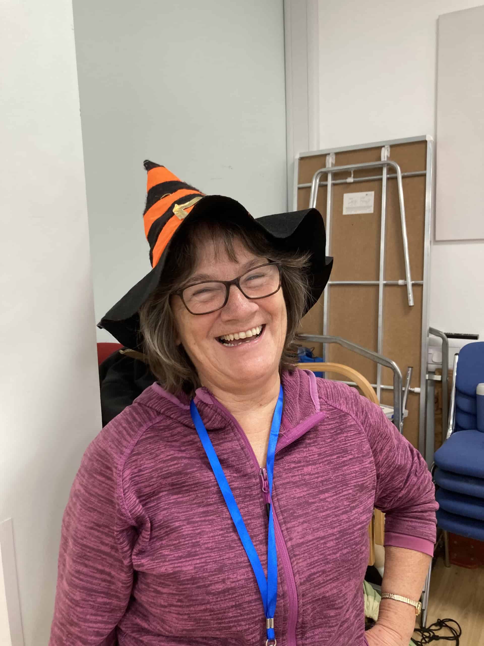 A smiling woman wearing glasses, a purple zip-up jumper, a blue lanyard, and a black and orange witch hat stands indoors in a room with chairs and folded tables in the background.