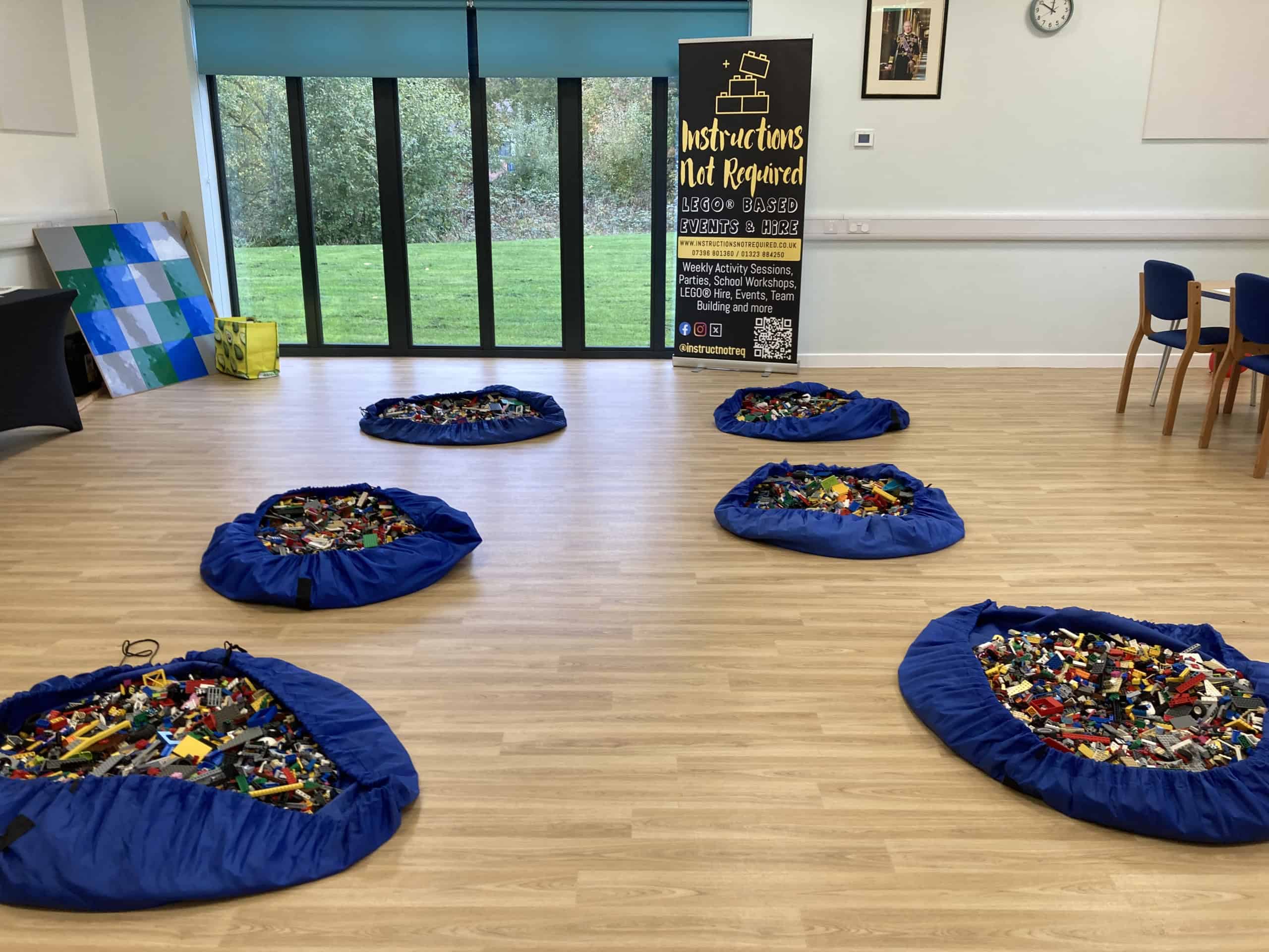 A spacious room with five blue mats filled with colourful Lego bricks on a wooden floor, chairs along the wall, large windows with a view of greenery, and a vertical sign at the back.