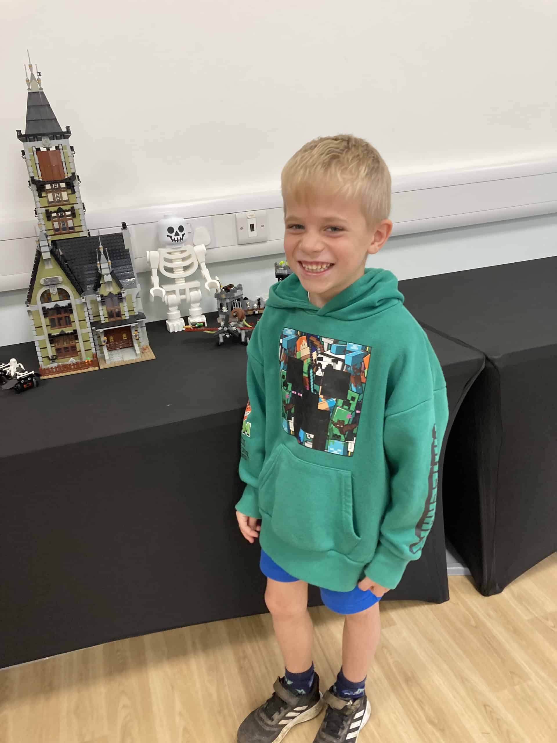 Young boy in a green hoodie smiles whilst standing next to a table displaying assembled LEGO sets, including a tall haunted house and a large white skeleton figure, in a bright indoor setting.