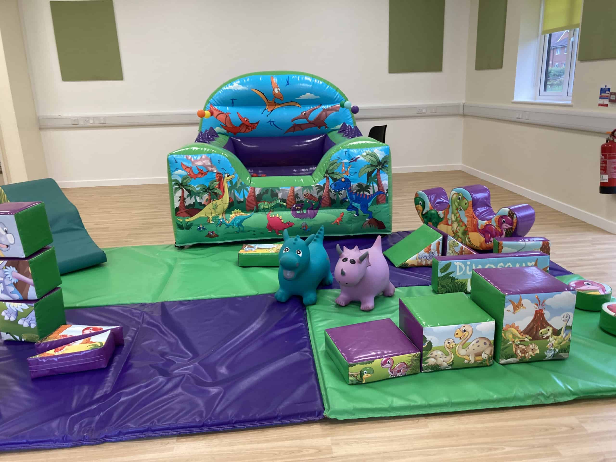 A colourful indoor play area with dinosaur-themed inflatable toys, foam blocks, soft mats, and two ride-on animal toys on a wooden floor in a bright room.