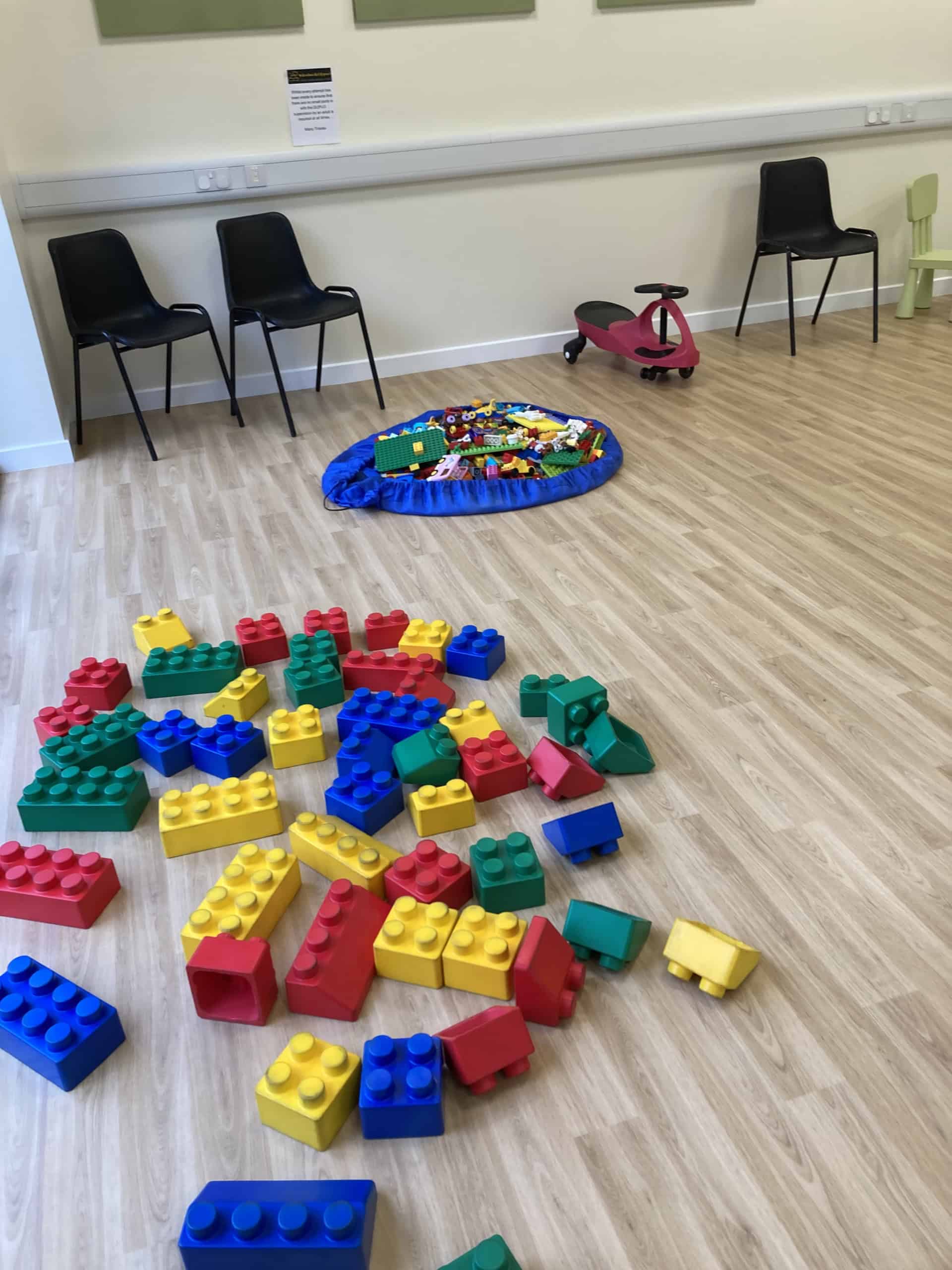 A playroom with large colourful plastic building blocks scattered on the floor, a pile of smaller toys on a blue mat, two black chairs, a green child’s chair, and a red ride-on toy in the background.