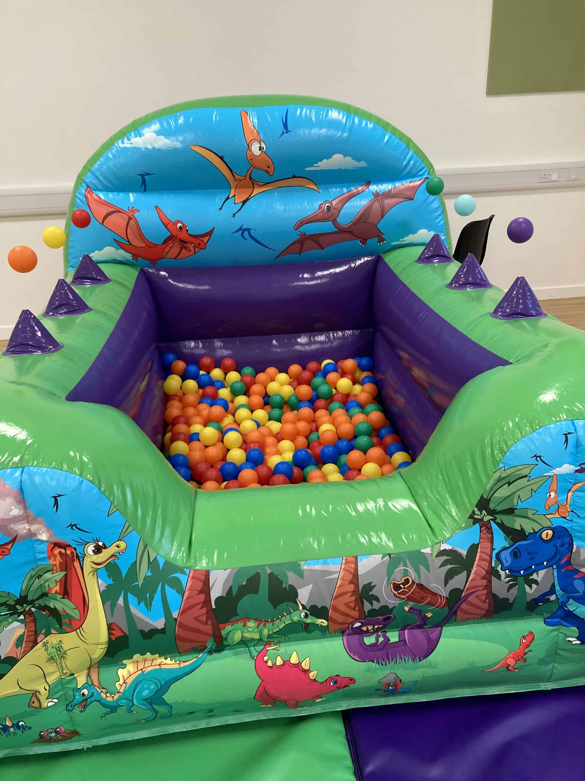 A colourful inflatable ball pit with dinosaur illustrations on the sides, filled with multicoloured plastic balls. The background shows part of a white wall and green mat on the floor.