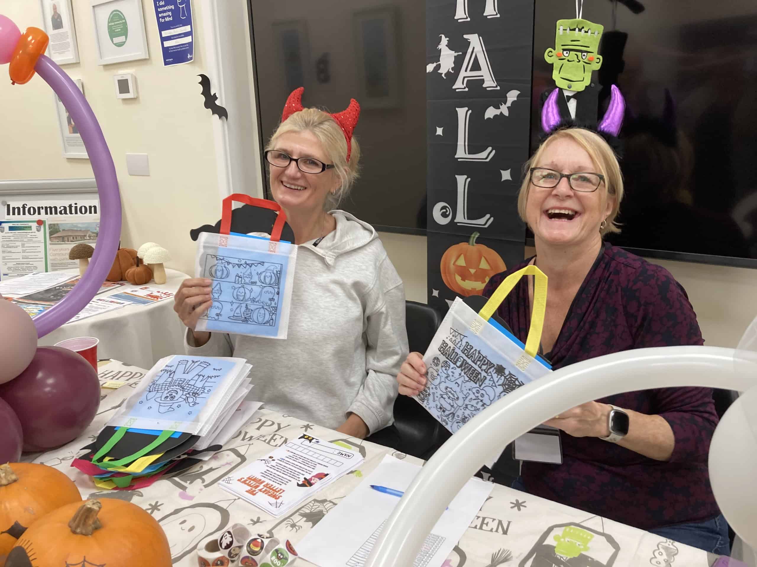 Two women wearing Halloween headbands and smiling at a decorated table hold up Halloween-themed activity bags. Pumpkins, balloons, and spooky decorations are on the table, creating a festive atmosphere.