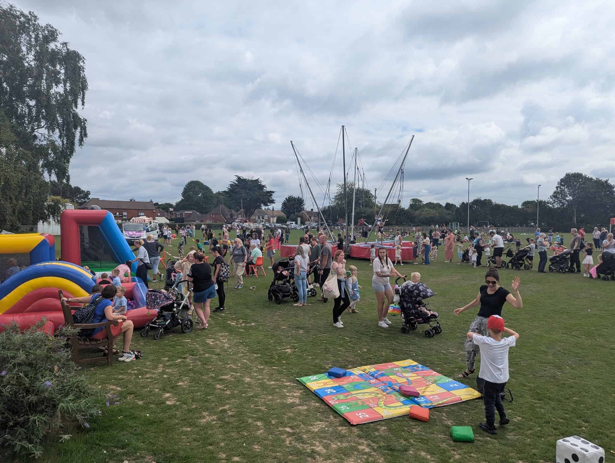 A busy outdoor fete with families and children enjoying activities like a bouncy castle, games, and rides on a grassy field under a cloudy sky. Pushchairs and colourful play mats are scattered around.